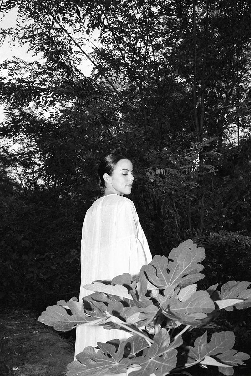 A woman in a flowing white dress stands gracefully amidst lush greenery